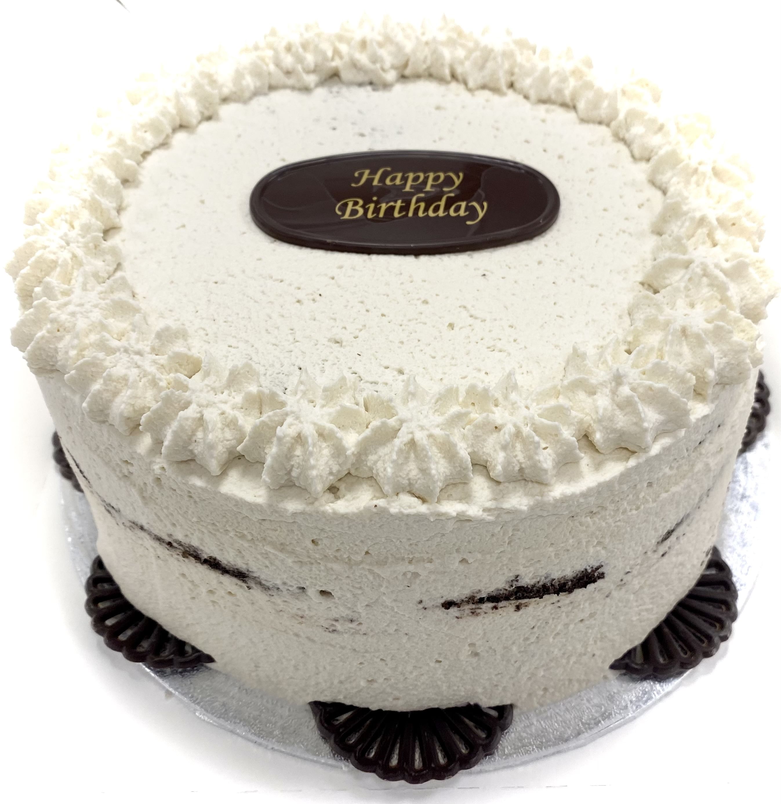 A round, frosted Black Forest gateaux with white icing, decorated with a scalloped edge of piped cream. The cake exudes chocolate goodness with accents around the bottom and a chocolate oval plaque on top that reads 'Happy Birthday' in golden letters. It hints at cherry liqueur for added richness.